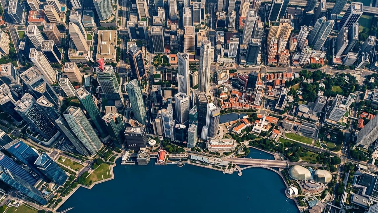 Editorial wide-angle aerial photograph of Singapore's central business district at mid-morning, with the towers of Raffles Place and the curved roofline of the Marina Bay financial centre catching hard equatorial sunlight against a deep tropical sky.