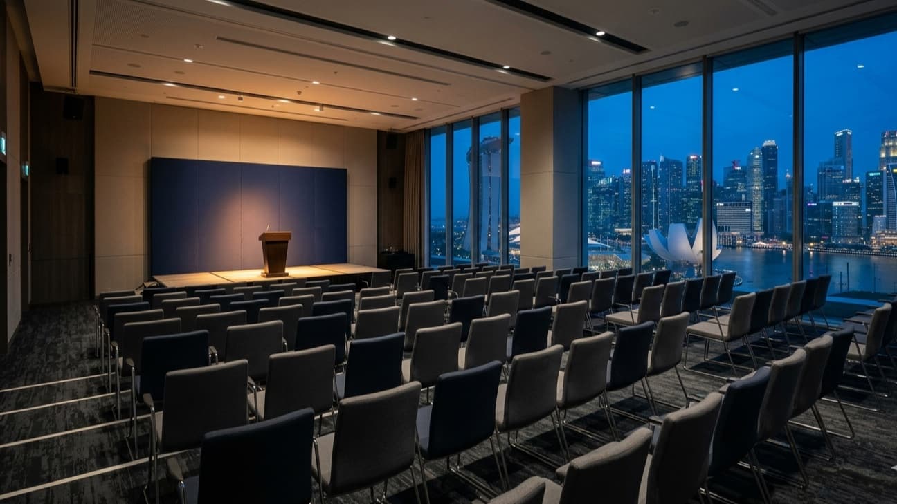 Editorial photograph of an empty conference auditorium in a Singapore financial-district hotel at pre-dawn blue hour, with rows of unoccupied chairs facing an institutional lectern lit warmly, and floor-to-ceiling windows revealing the Marina Bay skyline beyond.