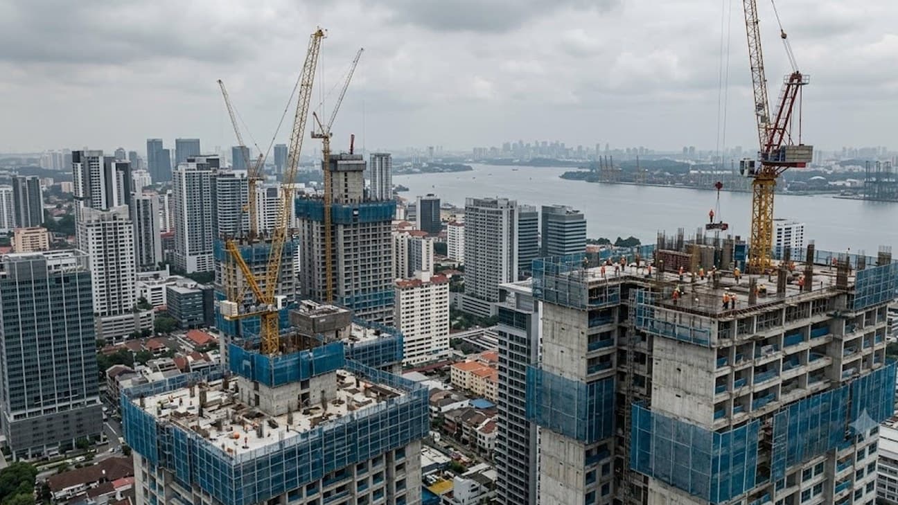 Aerial editorial photograph looking across the Johor Bahru skyline on an overcast midday, with two tower cranes rising over a cluster of half-built and completed high-rises wrapped in blue safety netting, small crews of construction workers visible on the rooftops, and the Straits of Johor and a hazy Singapore skyline in the distance.