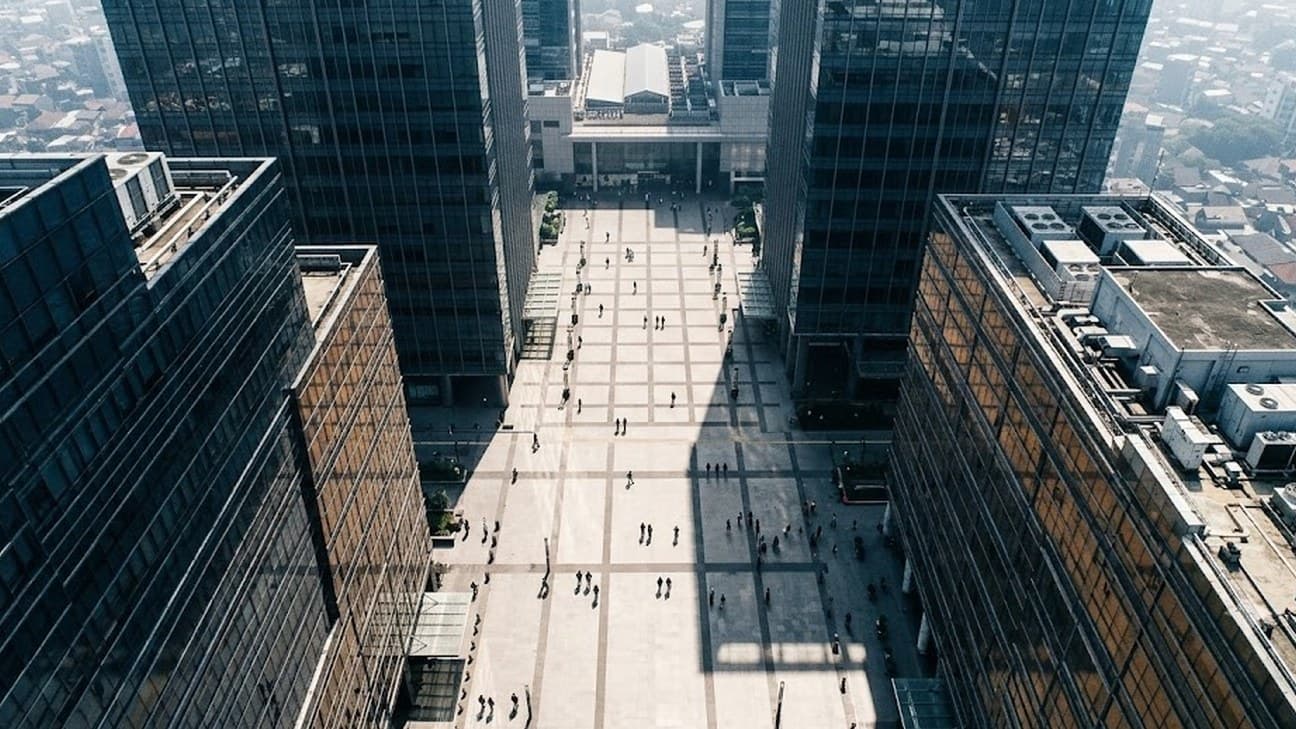 Wide aerial editorial photograph looking straight down on a Jakarta financial-district plaza at hard midday light, glass towers casting sharp shadows and pedestrians reduced to small distant figures crossing below.