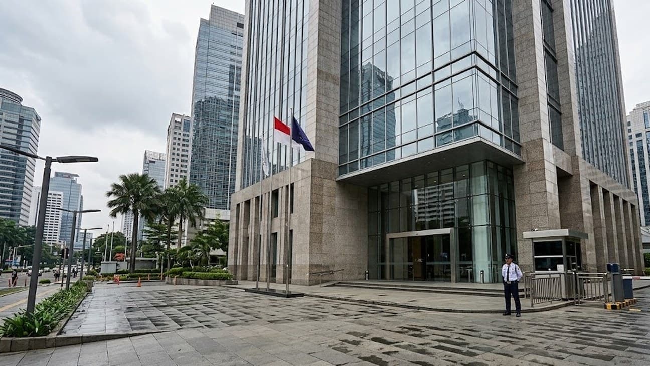 Wide editorial photograph of the exterior of a modern financial-district office tower in Southeast Asia under overcast midday light, glass-and-granite facade, palm-lined forecourt, and a single uniformed security guard standing near the entrance checkpoint.