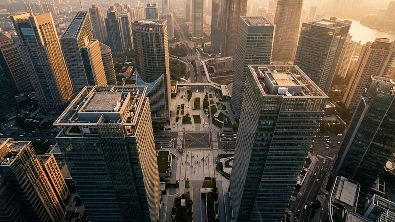 High-angle aerial editorial photograph of a Southeast Asian financial district at golden hour, glass-and-steel towers catching warm low sunlight, a plaza and crosswalk far below with tiny anonymous pedestrians crossing between buildings, long shadows stretching across the grid.
