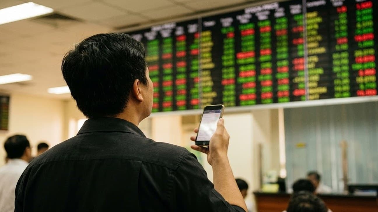 Photorealistic photograph of an investor viewed from behind, checking a smartphone while looking up at a large electronic stock ticker board covered in green and red price movements inside a Vietnamese brokerage investor centre.