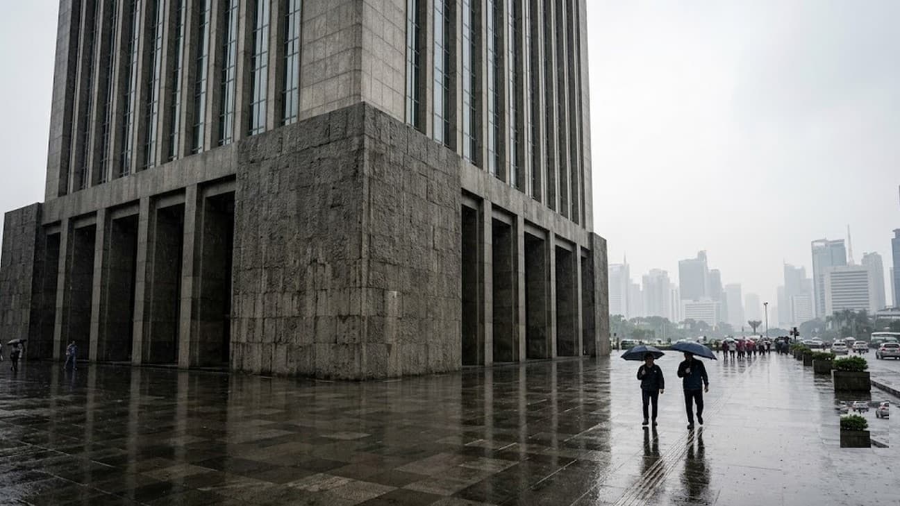 Two pedestrians with umbrellas cross a rain-slicked plaza beneath a monumental financial-regulator tower in Jakarta, hazy business-district skyline behind