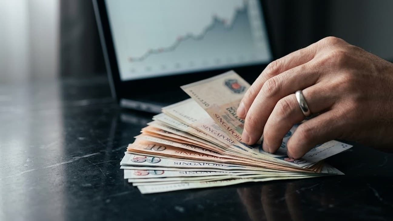 A close-up editorial photograph of an anonymous hand fanning a stack of Singapore dollar banknotes on a dark marble desk, with a blurred laptop screen showing an ascending funding chart in the background.