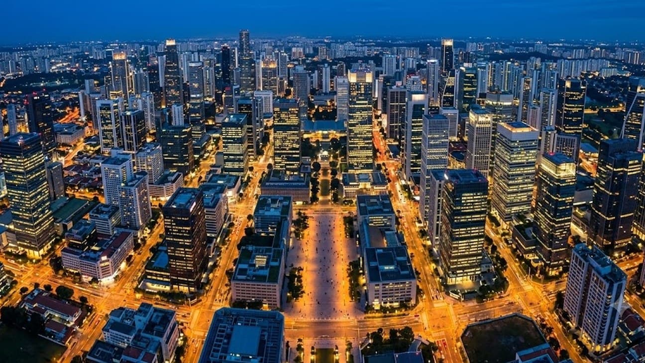 A wide editorial aerial photograph of a financial-district city grid at blue hour, illuminated office towers surrounding a lit central plaza, no legible text or logos.