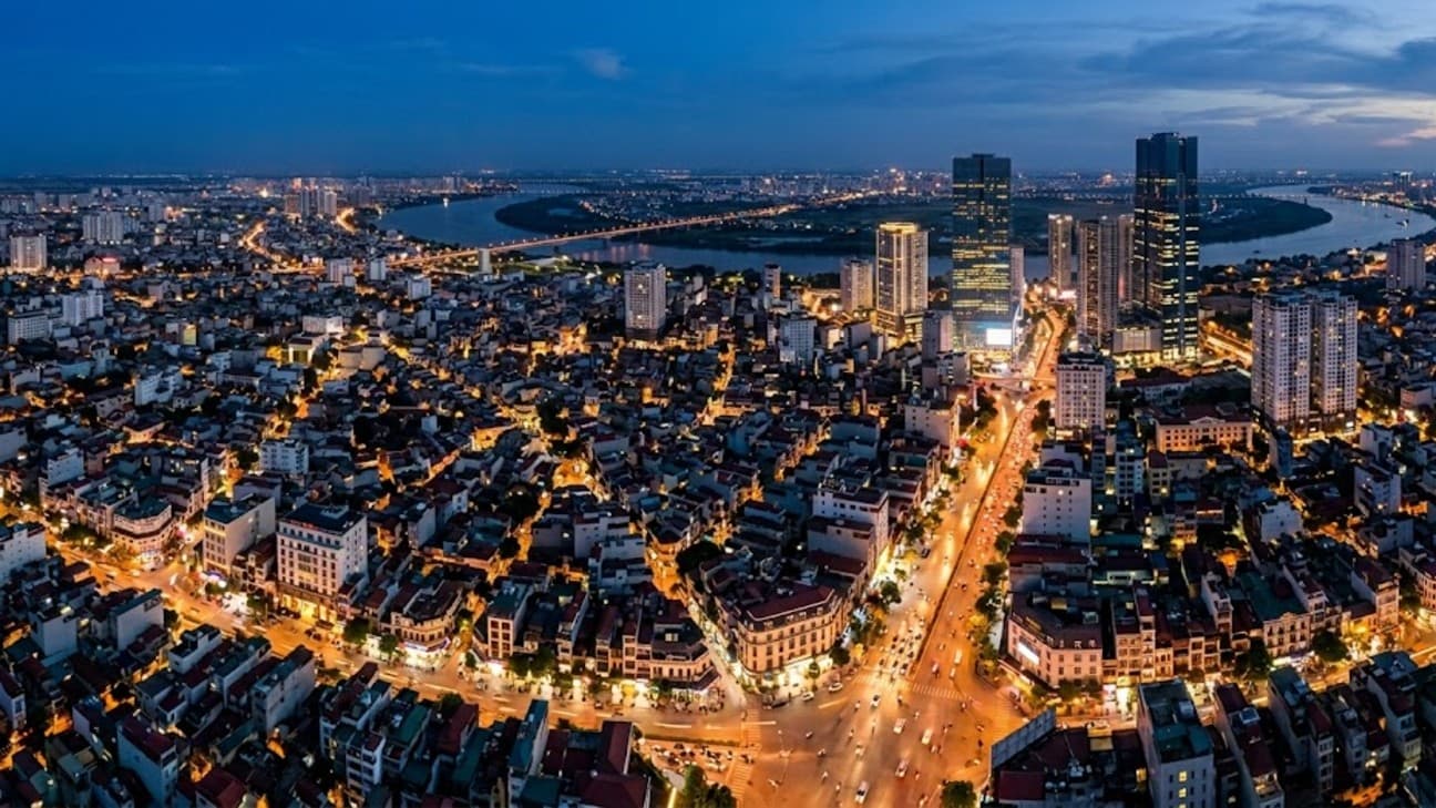 An aerial view of central Hanoi at blue hour, a dense grid of lit streets and mid-rise blocks with a cluster of modern towers and a river bend in the distance.