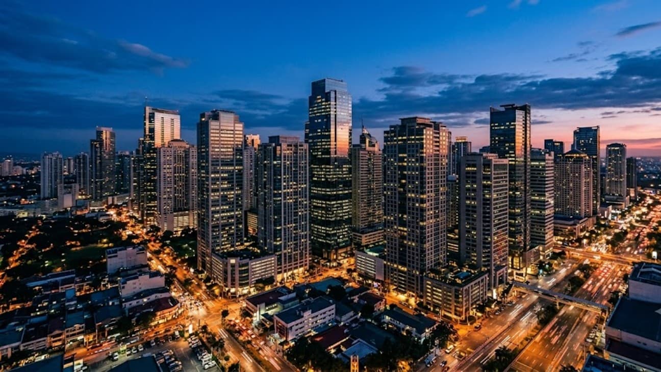 The Makati central business district skyline in Manila at dusk, clusters of lit glass office towers against a deep blue and orange evening sky.