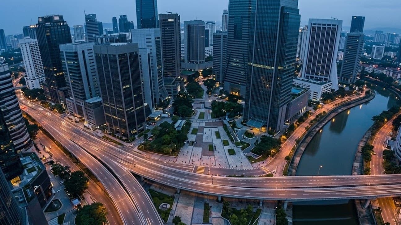 Aerial view of Kuala Lumpur's financial district at pre-dawn blue hour, with glass office towers, a paved plaza, an elevated road curving across the frame and a river bend lit by amber street lamps.