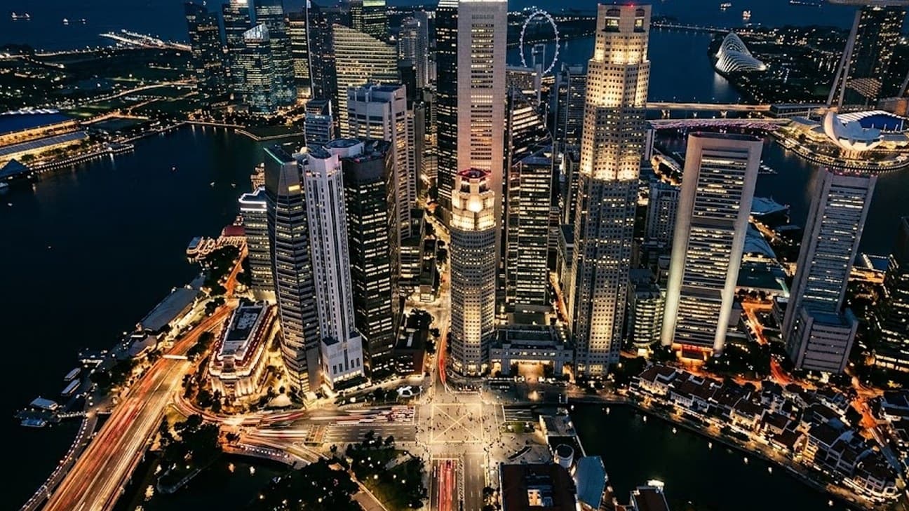 Photorealistic aerial photograph directly overhead of Singapore's Marina Bay financial district skyline at night, illuminated skyscraper office windows forming a lit grid pattern against dark water, long-exposure light trails from cars on the roads below, and tiny anonymous pedestrians crossing a plaza far below.
