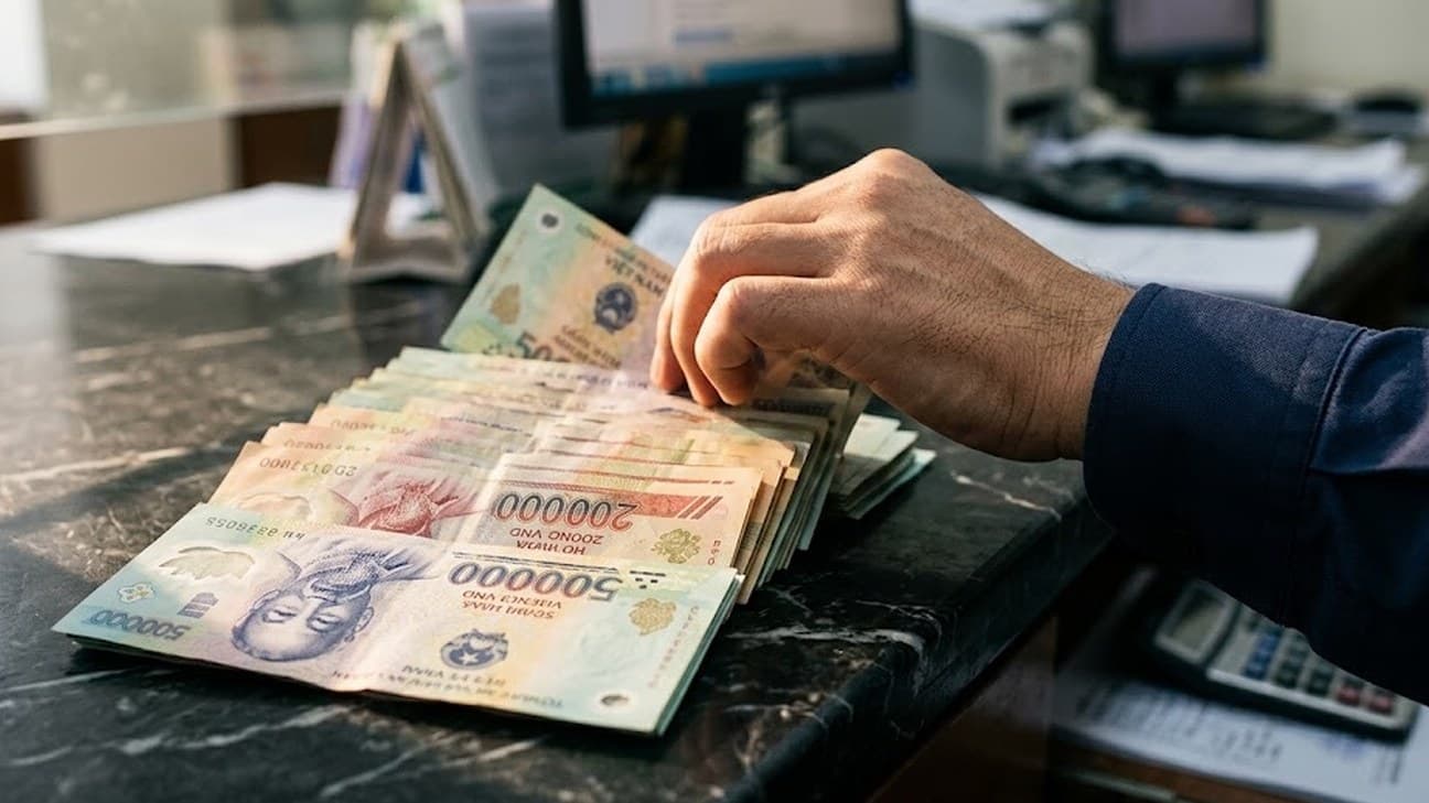 Photorealistic close-up editorial photograph of an anonymous hand counting a fanned stack of Vietnamese dong banknotes on a dark marble counter, with an out-of-focus office desk, monitors and a calculator in the background.