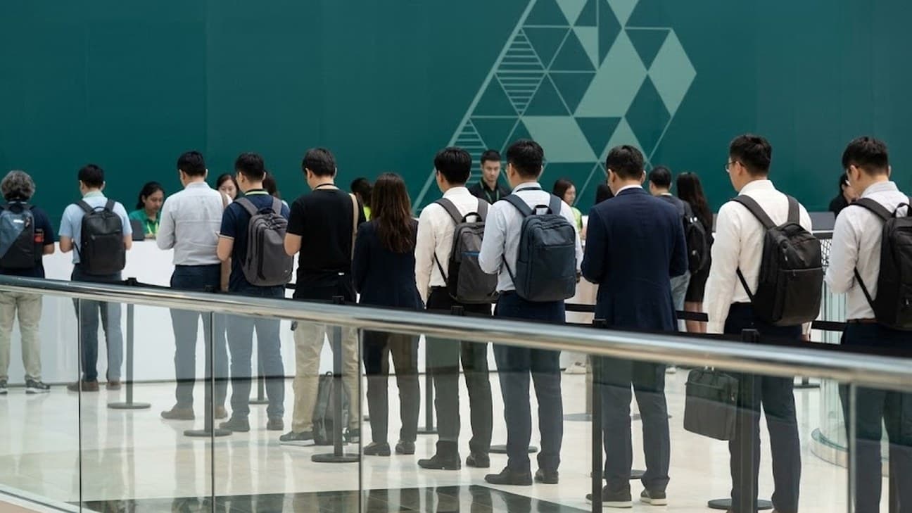 Photorealistic wide shot of a queue of conference attendees carrying backpacks, photographed from behind across a glass handrail, waiting to check in at a technology conference registration area with a teal geometric backdrop.