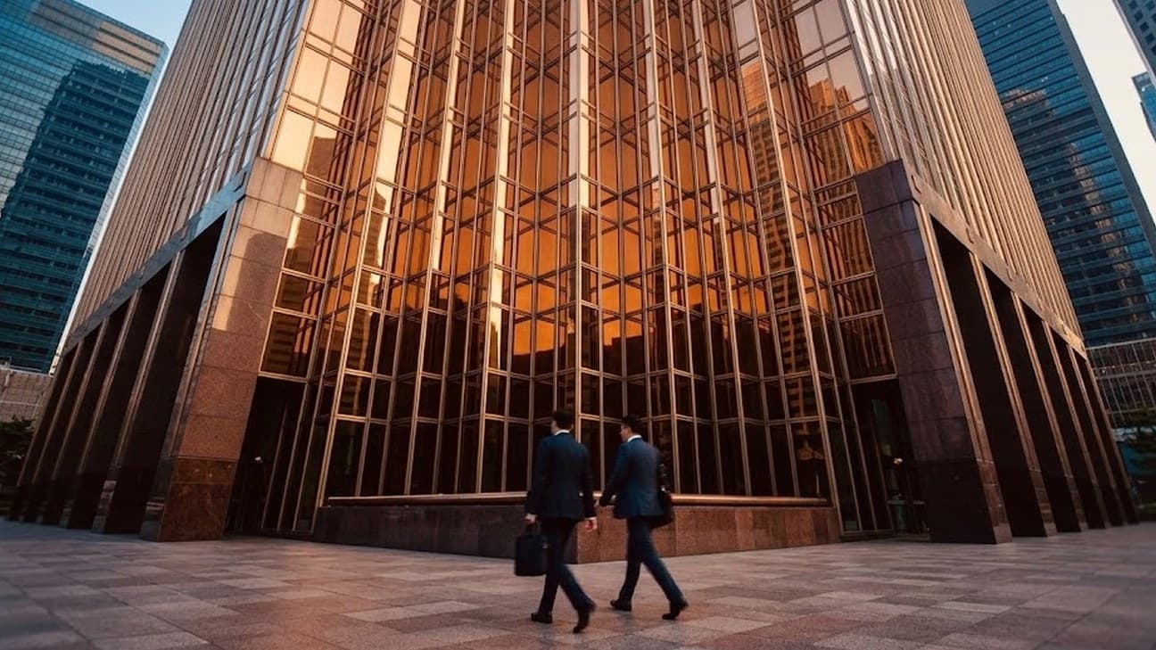Two office workers in business attire walk across a plaza in front of a glass-and-granite office tower in Singapore's financial district at golden hour.