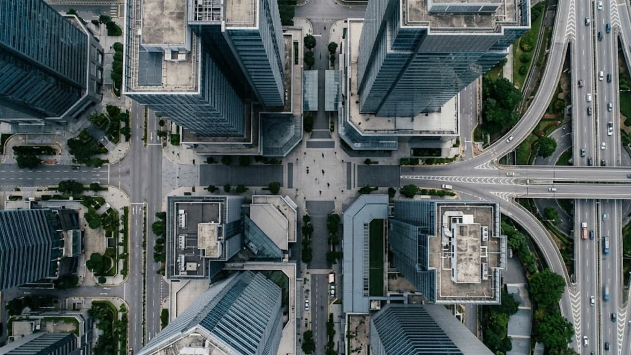 A wide editorial photograph looking straight down over the geometry of a Kuala Lumpur financial-district city block at overcast midday, towers and a highway interchange framing the grid; a few pedestrians appear as tiny distant figures crossing a plaza far below, no legible text or brand marks.