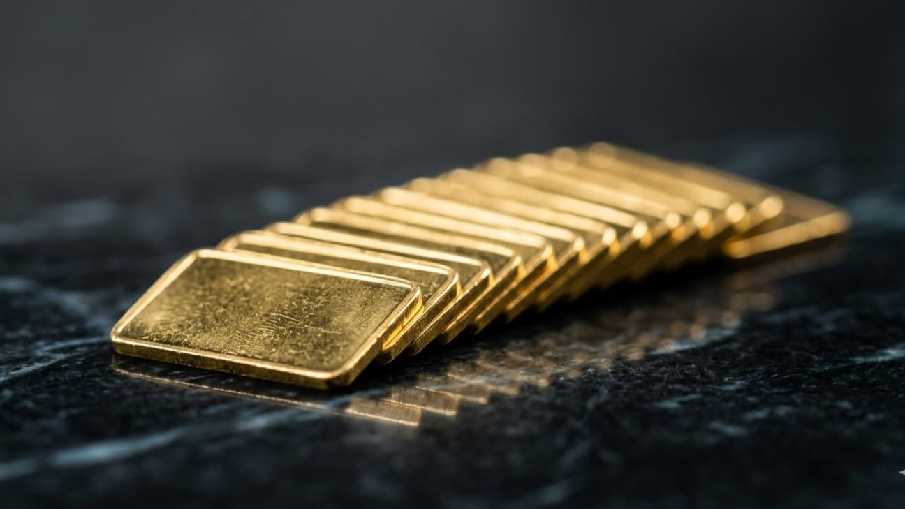 A fanned stack of one-gram gold bars rests on a dark polished surface under a single hard overhead light, with a faint reflection beneath, shot in the style of an editorial wire photograph.